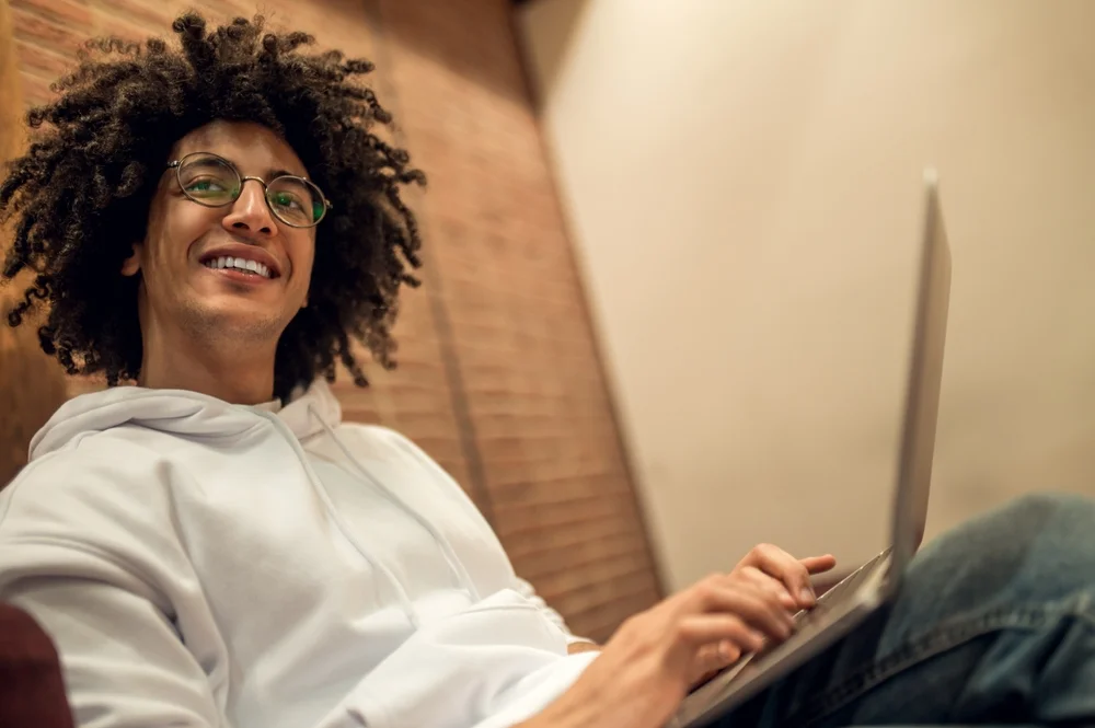Estudante sorrindo e prestando atenção na tela de um notebook enquanto está sentado confortavelmente em um sofá, ilustrando a praticidade e a autonomia de realizar uma Formação Continuada no formato 100% online.